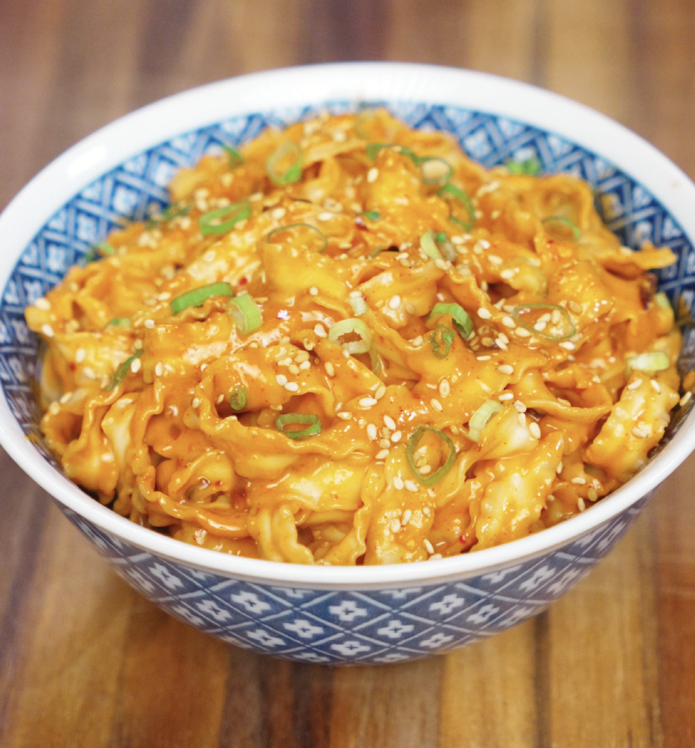 A bowl brimming with glossy gochujang peanut noodles, topped with scallions and sesame seeds. The sauce is rich and orange-red, coating every curl of noodle with bold, nutty flavor.