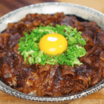 A bowl of rice topped with caramelized yakiniku beef, sliced scallions, and a raw egg yolk in the center. The glossy glaze on the beef adds a rich, savory shine that contrasts with the bright yellow yolk.