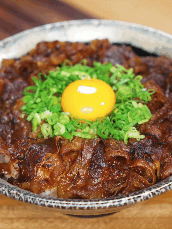 A bowl of rice topped with caramelized yakiniku beef, sliced scallions, and a raw egg yolk in the center. The glossy glaze on the beef adds a rich, savory shine that contrasts with the bright yellow yolk.