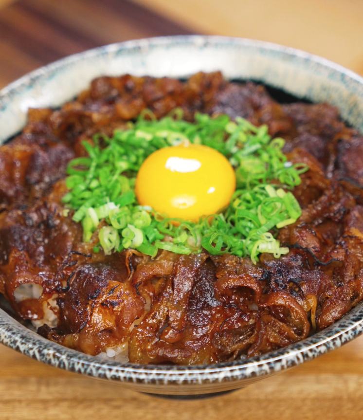 A bowl of rice topped with caramelized yakiniku beef, sliced scallions, and a raw egg yolk in the center. The glossy glaze on the beef adds a rich, savory shine that contrasts with the bright yellow yolk.