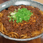 A bowl of garlic beef noodles topped with crispy garlic and chopped scallions. The glossy noodles glisten under the light, ready to eat.