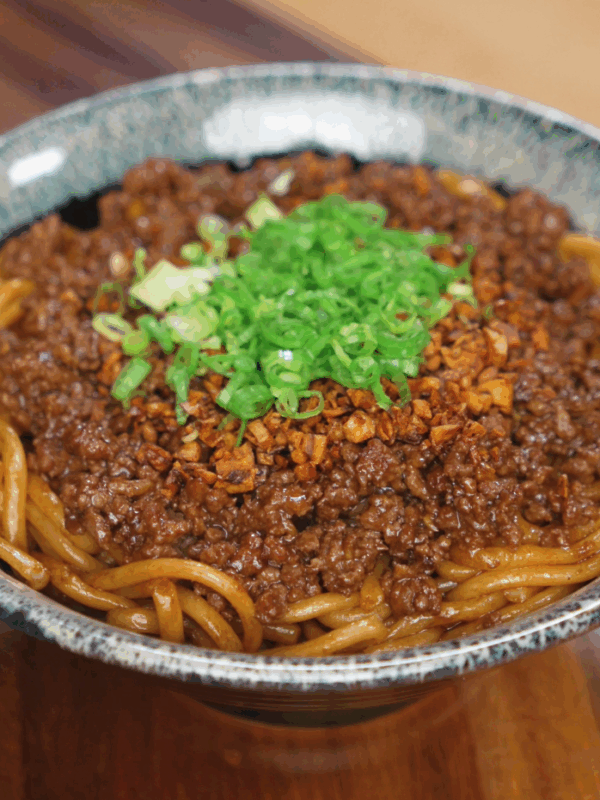 A bowl of garlic beef noodles topped with crispy garlic and chopped scallions. The glossy noodles glisten under the light, ready to eat.