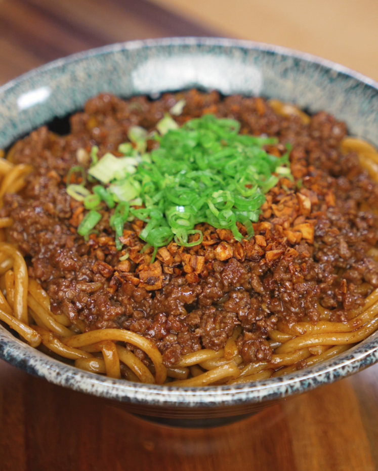 A bowl of garlic beef noodles topped with crispy garlic and chopped scallions. The glossy noodles glisten under the light, ready to eat.