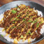 A completed bowl of Japanese BBQ beef served over rice, topped with a drizzle of sriracha mayo, sesame seeds, and chopped scallions. The beef is deeply browned and caramelized, contrasting beautifully with the white rice and vibrant garnishes.