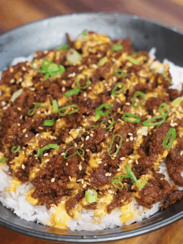 A completed bowl of Japanese BBQ beef served over rice, topped with a drizzle of sriracha mayo, sesame seeds, and chopped scallions. The beef is deeply browned and caramelized, contrasting beautifully with the white rice and vibrant garnishes.