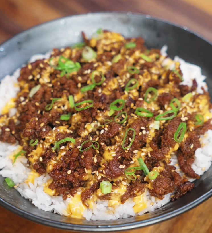 A completed bowl of Japanese BBQ beef served over rice, topped with a drizzle of sriracha mayo, sesame seeds, and chopped scallions. The beef is deeply browned and caramelized, contrasting beautifully with the white rice and vibrant garnishes.
