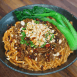 A bowl of spicy beef noodles topped with savory ground beef, chili oil, crushed peanuts, chopped scallions, and vibrant green bok choy. The thick sauce coats the noodles, making the dish look rich, hearty, and full of umami.