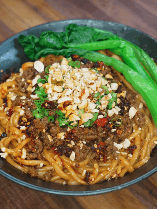 A bowl of spicy beef noodles topped with savory ground beef, chili oil, crushed peanuts, chopped scallions, and vibrant green bok choy. The thick sauce coats the noodles, making the dish look rich, hearty, and full of umami.