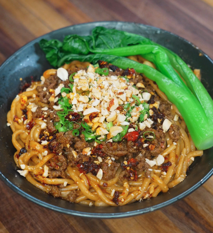 A bowl of spicy beef noodles topped with savory ground beef, chili oil, crushed peanuts, chopped scallions, and vibrant green bok choy. The thick sauce coats the noodles, making the dish look rich, hearty, and full of umami.