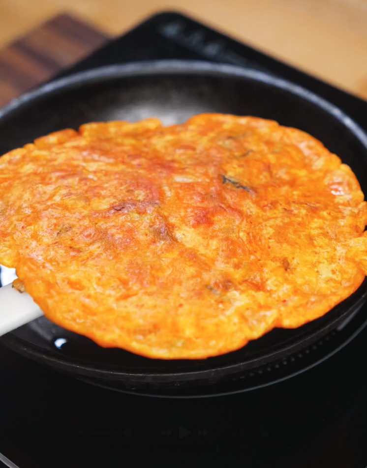 A golden-brown kimchi pancake is being flipped with a spatula inside a cast iron pan. The surface is evenly crisped, indicating it’s perfectly cooked on one side.