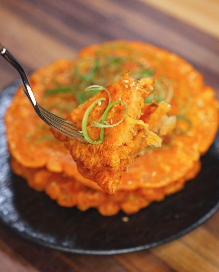 A fork lifts a crispy triangle of kimchi pancake from the stack, showcasing the pancake’s golden brown edges, savory filling, and topping of curly scallions and sesame seeds.