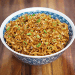 A blue patterned bowl filled with glistening garlic butter noodles, coated in soy-based sauce and garnished with sliced scallions and toasted sesame seeds on a wooden table.