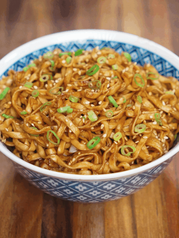 A blue patterned bowl filled with glistening garlic butter noodles, coated in soy-based sauce and garnished with sliced scallions and toasted sesame seeds on a wooden table.