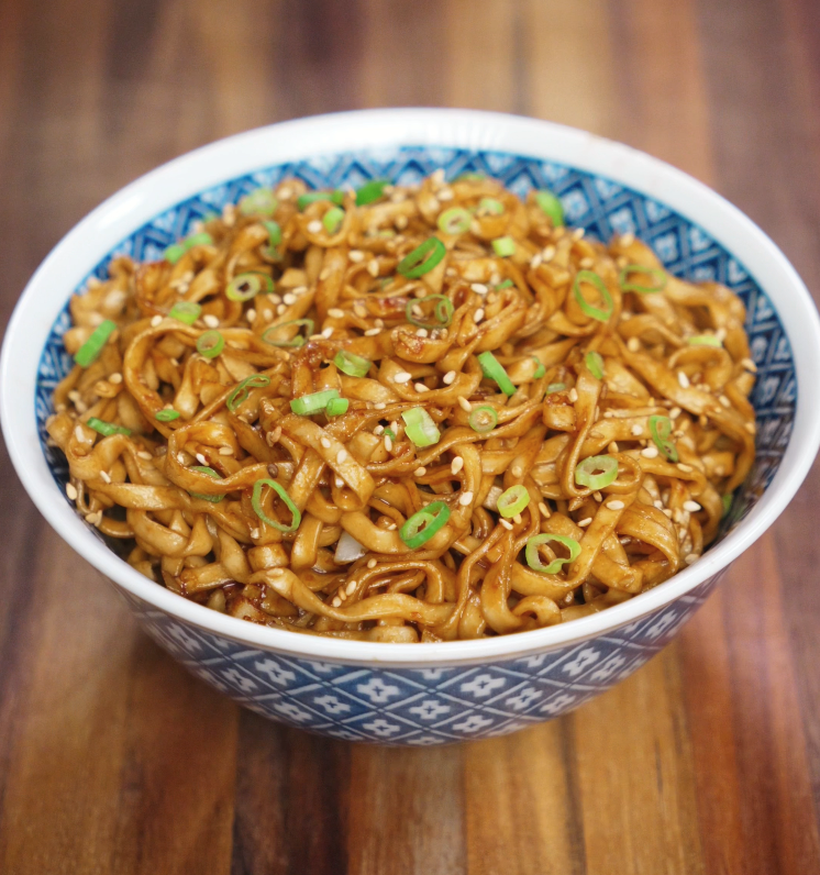 A blue patterned bowl filled with glistening garlic butter noodles, coated in soy-based sauce and garnished with sliced scallions and toasted sesame seeds on a wooden table.