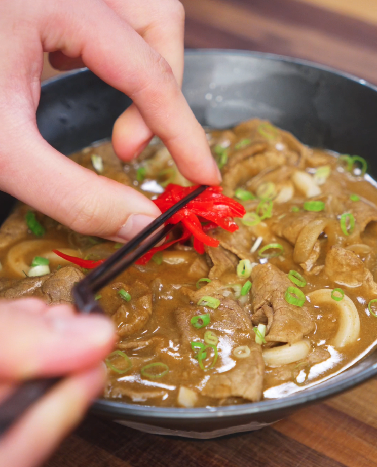 A hand uses chopsticks to gently place a bundle of bright red pickled ginger on top of the curry udon. The tangy garnish adds a sharp contrast to the savory curry base.