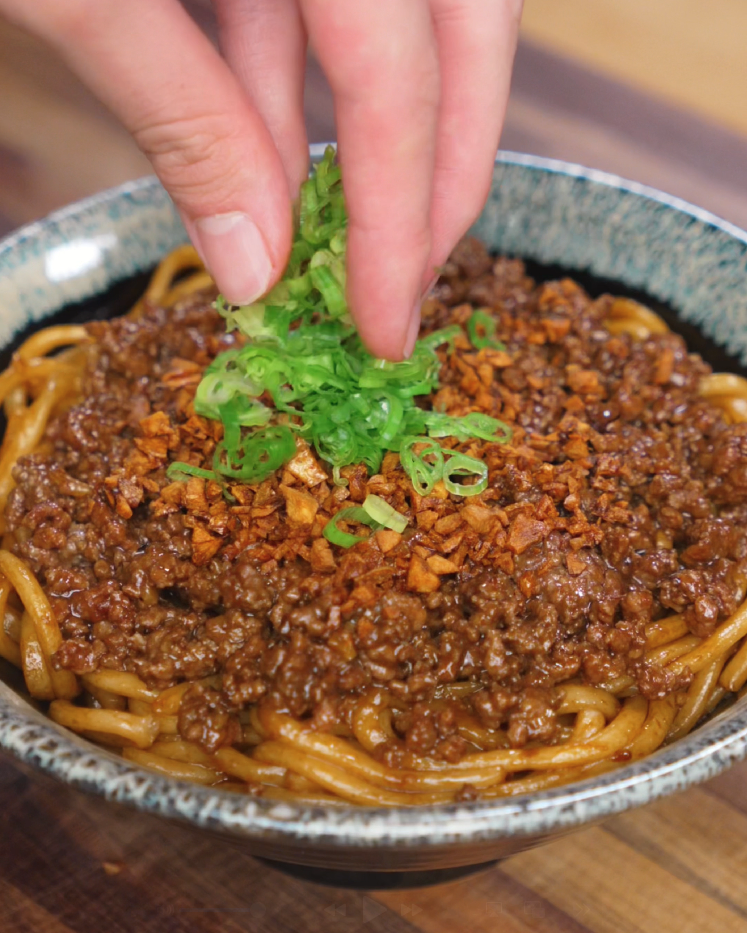 A hand sprinkles fresh green scallions over a bowl of garlic beef noodles, topped with crispy garlic and saucy ground beef. The vibrant garnish adds color and freshness to the rich, savory dish.