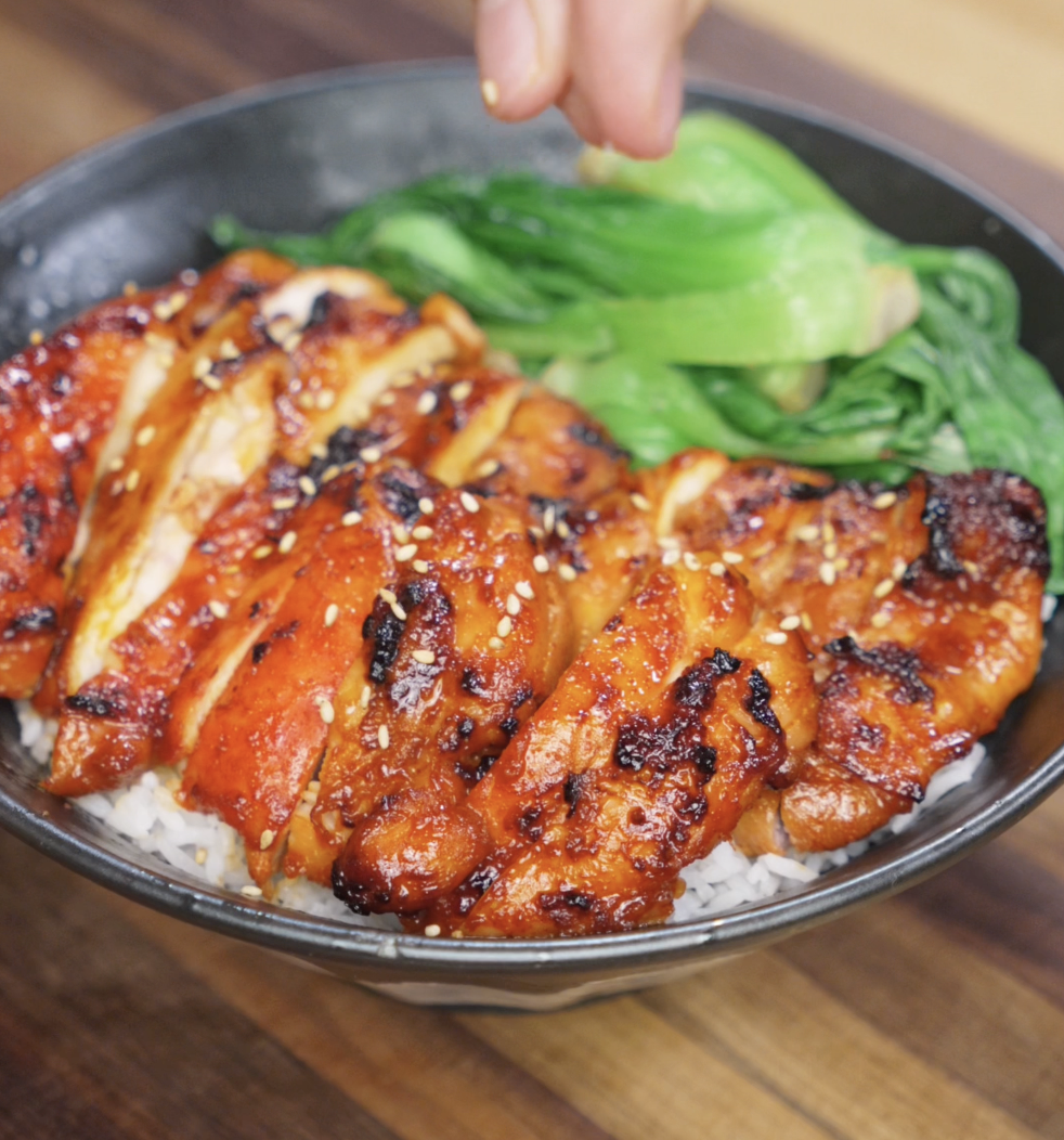 A hand sprinkles sesame seeds over a finished gochujang chicken bowl. The vibrant red glaze, green bok choy, and toasted sesame toppings complete the dish with flavor and texture.