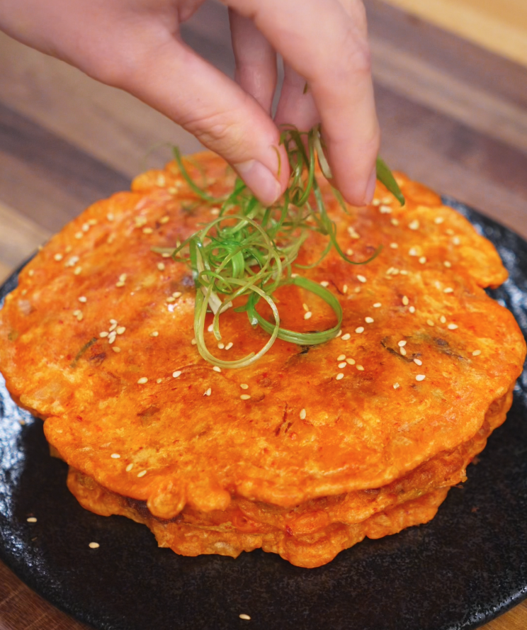 A hand places a pile of thinly sliced green onions atop a stack of kimchi pancakes. The pancakes are already sprinkled with sesame seeds, ready to be served hot and crispy.