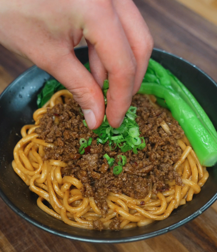 A hand sprinkles sliced green onions over a bowl of saucy beef noodles. The beef is deeply browned and savory, with tender bok choy adding freshness and contrast.