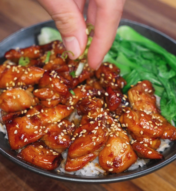 A hand sprinkles chopped scallions over a bowl of soy butter chicken glazed with sesame seeds, served over white rice with bok choy — the final garnish for color, crunch, and freshness.