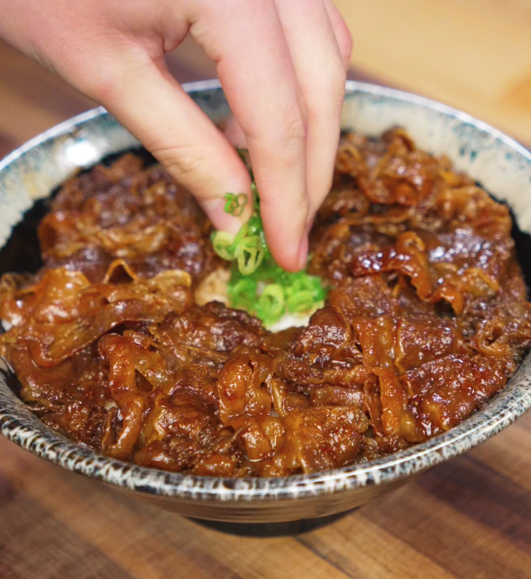 A hand places a small pile of freshly chopped scallions in the center of a yakiniku beef bowl. The greens sit atop a glossy bed of glazed beef, ready for an egg yolk topping.