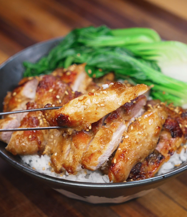 Chopsticks lift a juicy slice of lemon pepper chicken over the rice bowl. The chicken is golden, glossy, and flecked with sesame seeds, ready to eat.