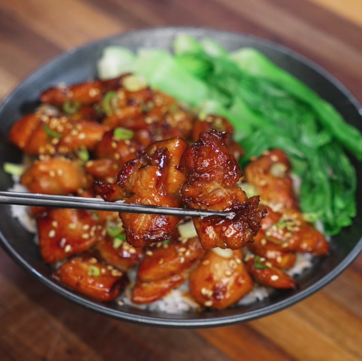 A glossy bite of soy butter chicken is lifted with chopsticks from a bowl filled with sesame-topped chicken, steamed rice, and sautéed bok choy, showcasing its sticky texture and caramelized exterior.