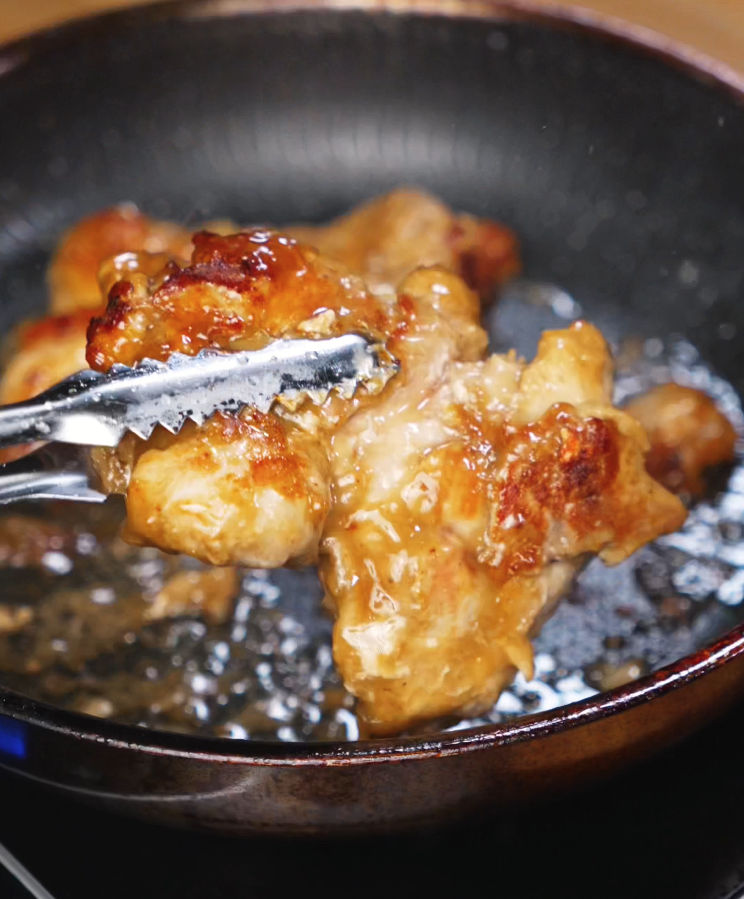 A pair of tongs lifts a seared chicken thigh from a skillet. The chicken is juicy and perfectly browned, with a light glaze from the lemon pepper marinade.