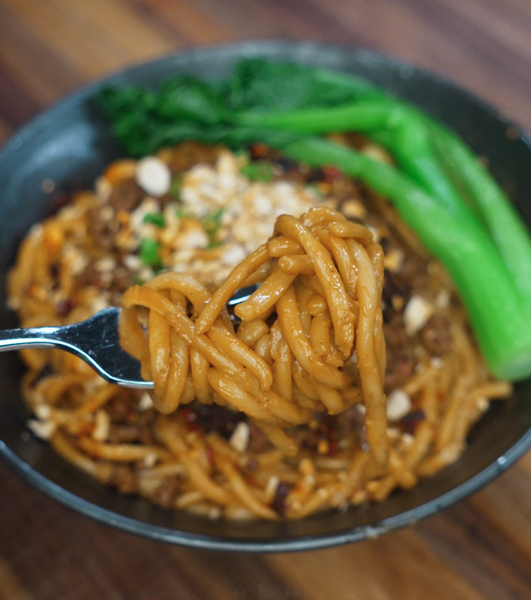 A fork lifts a tangle of thick, saucy noodles from the bowl, showing how well the spicy peanut sauce clings to the strands. The background reveals tender beef and crisp bok choy still in the bowl.