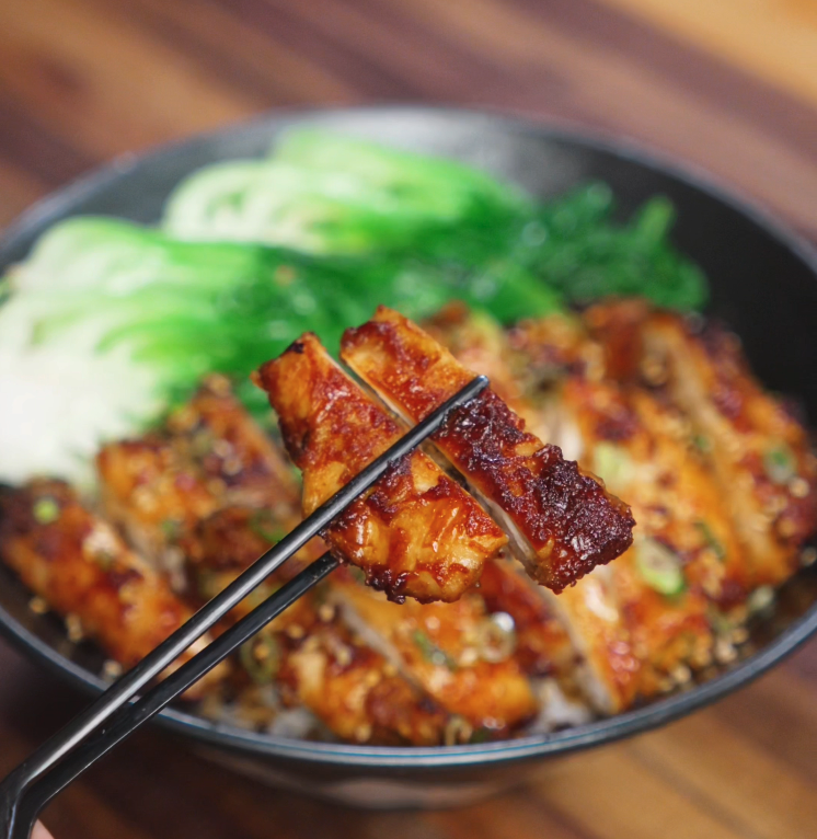 A pair of chopsticks holds up a juicy, golden-brown slice of peanut glazed chicken, highlighting the glossy, caramelized exterior. The bowl below is filled with rice, greens, and the remaining perfectly cooked chicken pieces.