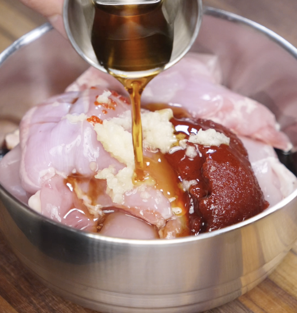 A close-up of gochujang, minced garlic, soy sauce, and sesame oil being poured over raw chicken thighs in a metal bowl. This spicy, sweet, and savory marinade forms the bold flavor base for the glazed chicken.