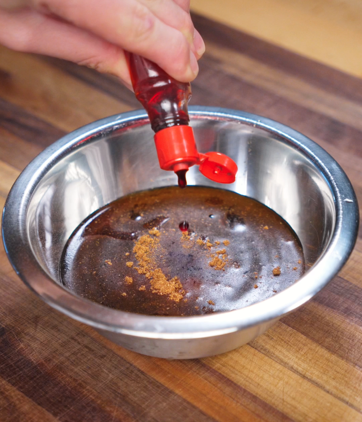 A hand squeezes red food coloring into a metal bowl filled with soy sauce, hoisin, honey, garlic, and five spice powder. This vibrant marinade creates the signature glossy red glaze of traditional char siu pork belly.