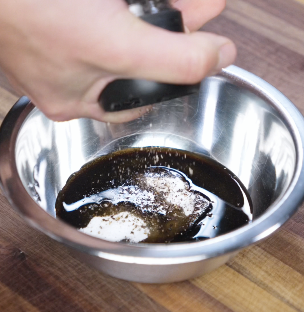 A hand adds freshly ground black pepper to a dark, glossy sauce mixture in a stainless steel bowl. The sauce combines soy sauces, oyster sauce, sugar, and pepper for a bold, umami-rich base.