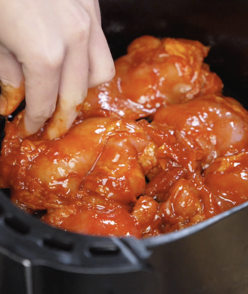 A hand arranges gochujang-marinated chicken thighs in an air fryer basket. The vibrant red coating glistens, ready to be crisped and caramelized in the air fryer.