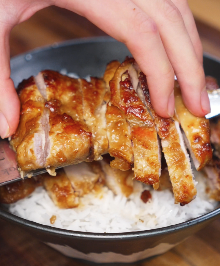 A hand carefully arranges sliced lemon pepper chicken over a bed of fluffy white rice using a knife. The glistening chicken contrasts with the soft rice underneath.