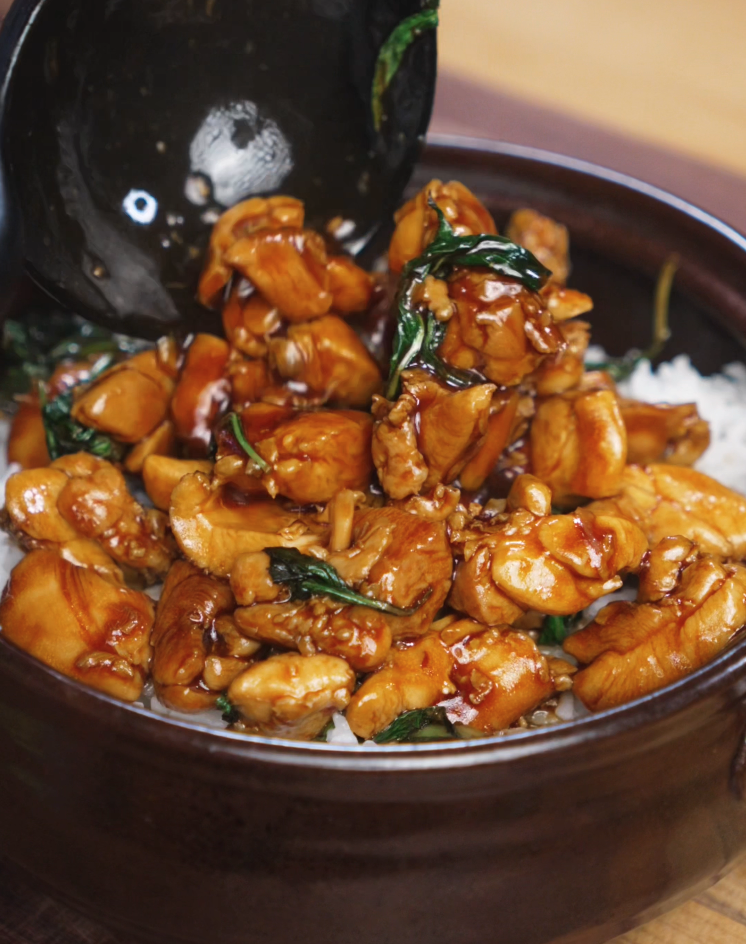 A generous spoonful of glossy Thai basil chicken is ladled over a bowl of white rice. The sauce clings to each piece of chicken and basil for a comforting, flavorful meal.