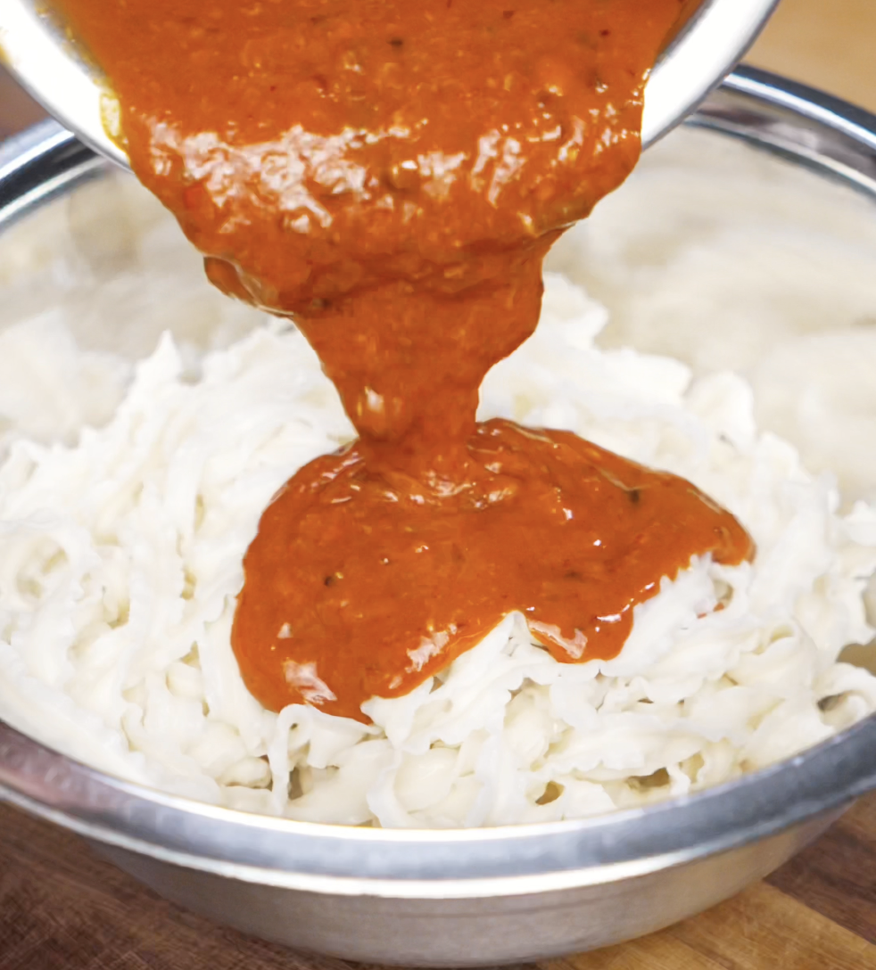 A stream of thick, fiery orange gochujang peanut sauce pours over a bowl of freshly cooked noodles. The velvety texture promises a creamy, spicy finish.