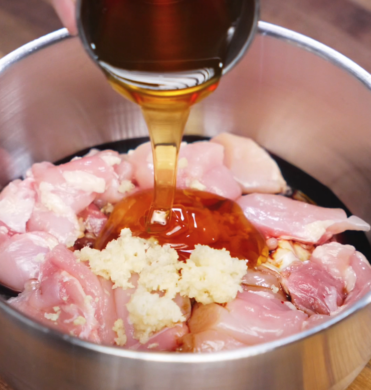 A stream of golden honey is poured over a bowl of raw chicken thighs mixed with minced garlic and soy sauce, highlighting the beginning of the soy butter chicken marinade.