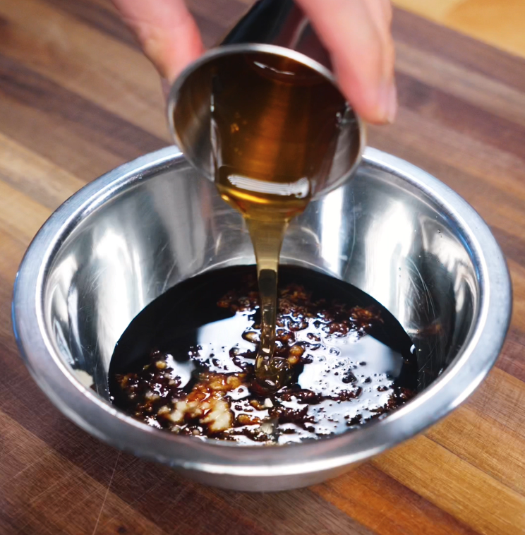 A golden stream of honey soy BBQ sauce is poured into a bowl containing minced garlic, grated ginger, and soy sauces. The mixture glistens with richness, ready to coat the beef with sweet and savory flavor.