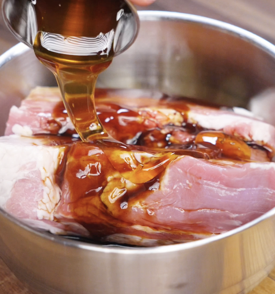 A metal cup pours a stream of glossy honey soy marinade over raw pork belly in a stainless steel bowl. The thick sauce coats the meat in preparation for a short marination.