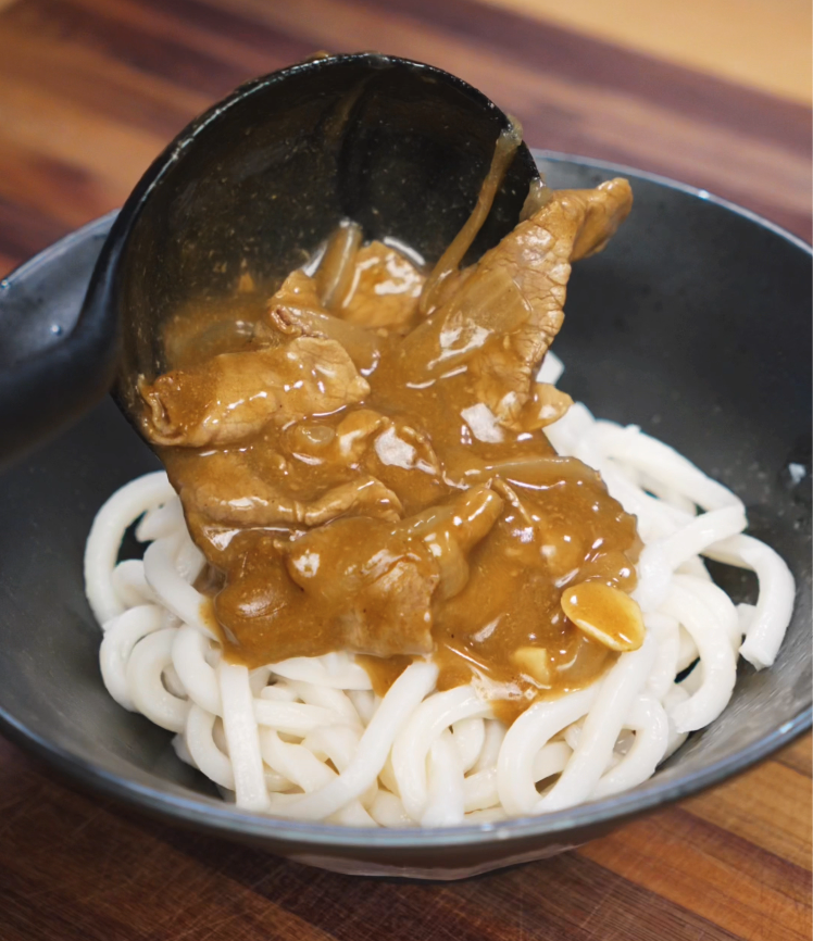 A ladle pours thick Japanese curry sauce with beef and onions over a bowl of chewy udon noodles. The savory gravy clings to each strand, creating a comforting noodle dish.