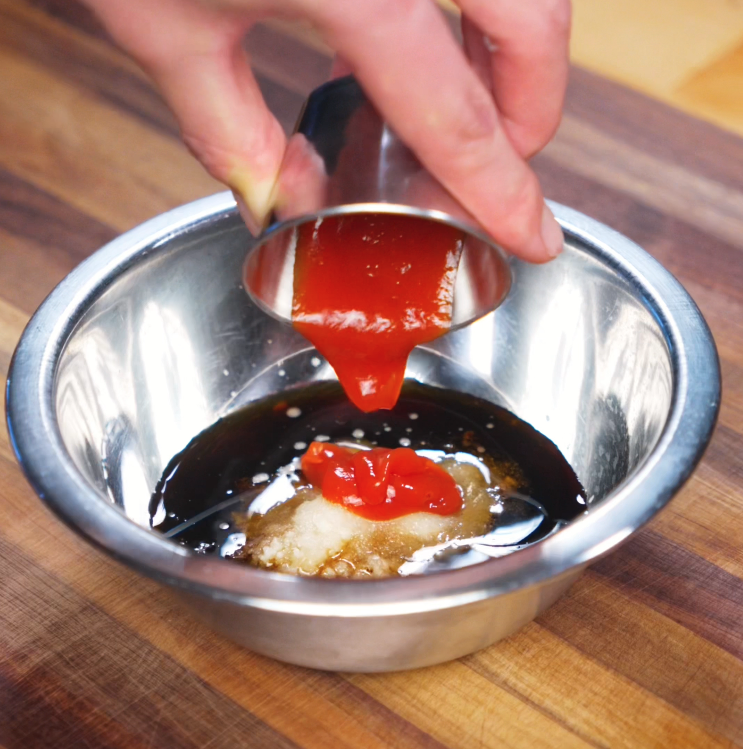 A metal sauce cup pours a stream of bright red ketchup into a bowl of soy sauce, sugar, and garlic. This forms the sweet-savory base of the yakiniku glaze.