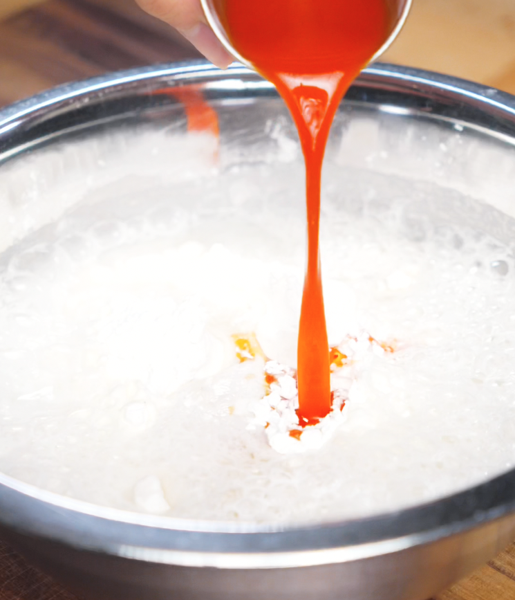 Bright red kimchi juice is being poured into a foamy batter mixture of sparkling water, flour, and cornstarch in a stainless steel bowl, adding color and bold flavor to the kimchi pancake base.