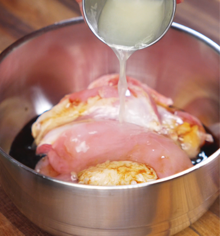 Fresh lemon juice is poured from a metal cup over raw chicken thighs sitting in soy sauce and garlic. This quick marinade adds brightness and tenderness before cooking.