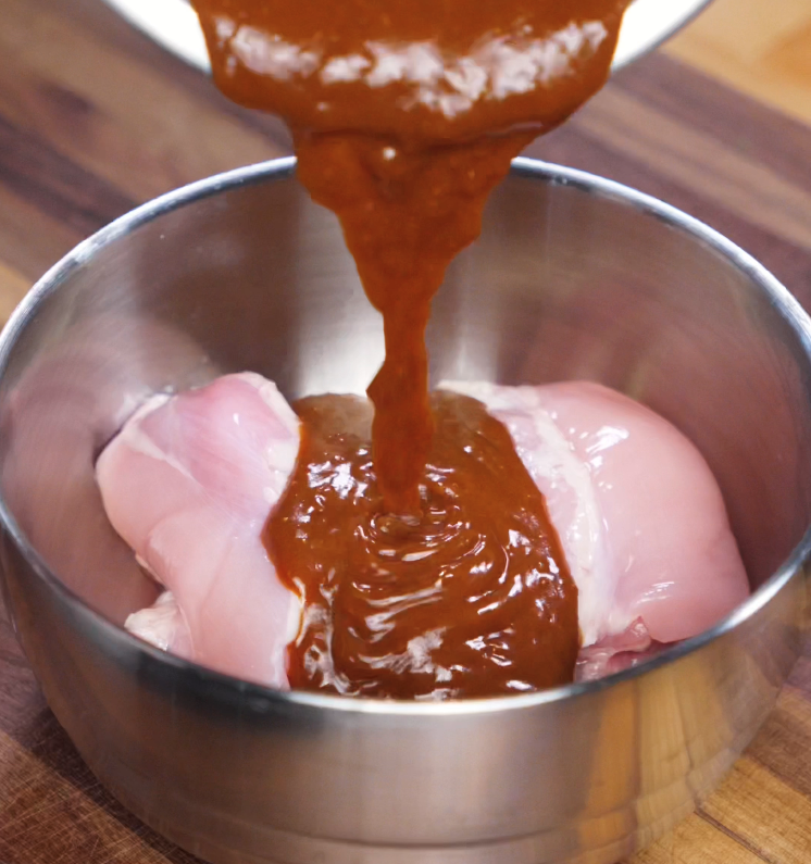 A thick, rich peanut marinade is being poured over raw boneless chicken thighs in a metal bowl. The sauce generously coats the chicken, signaling the start of a short but flavor-packed marination process.