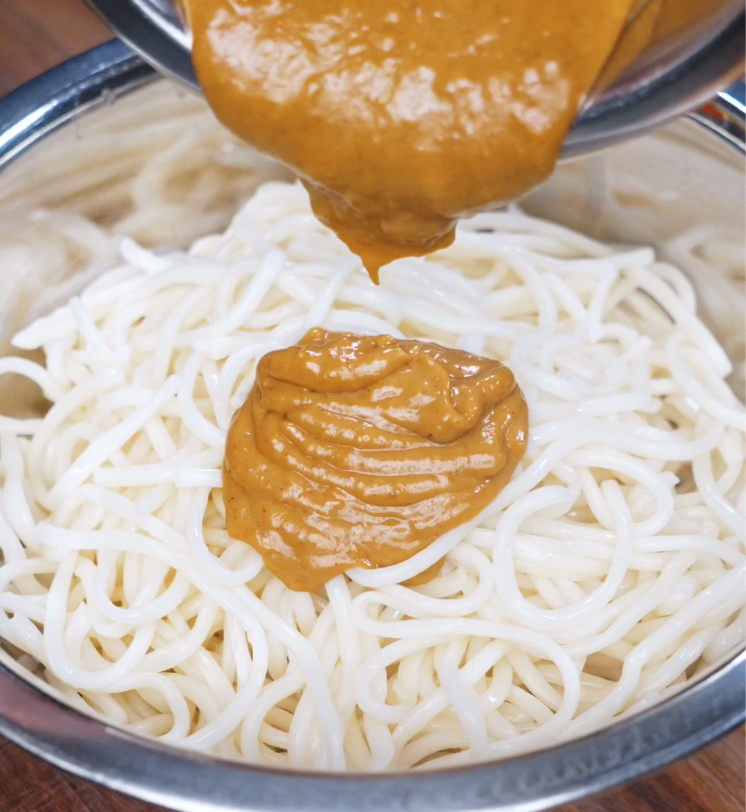 Thick, golden-brown peanut sauce is poured generously over freshly cooked noodles in a metal bowl. The creamy texture and glossy sheen signal a rich, flavorful base for the dish.