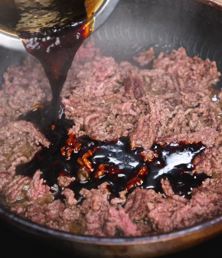 A rich, dark Japanese BBQ sauce is poured from a bowl onto partially cooked ground beef in a skillet. The sauce immediately begins to sizzle, enveloping the meat in a glossy coat of flavor.