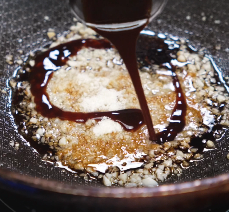 A thick stream of soy-based sauce is being poured into a pan of sizzling garlic butter, forming a rich and aromatic base for the noodles.