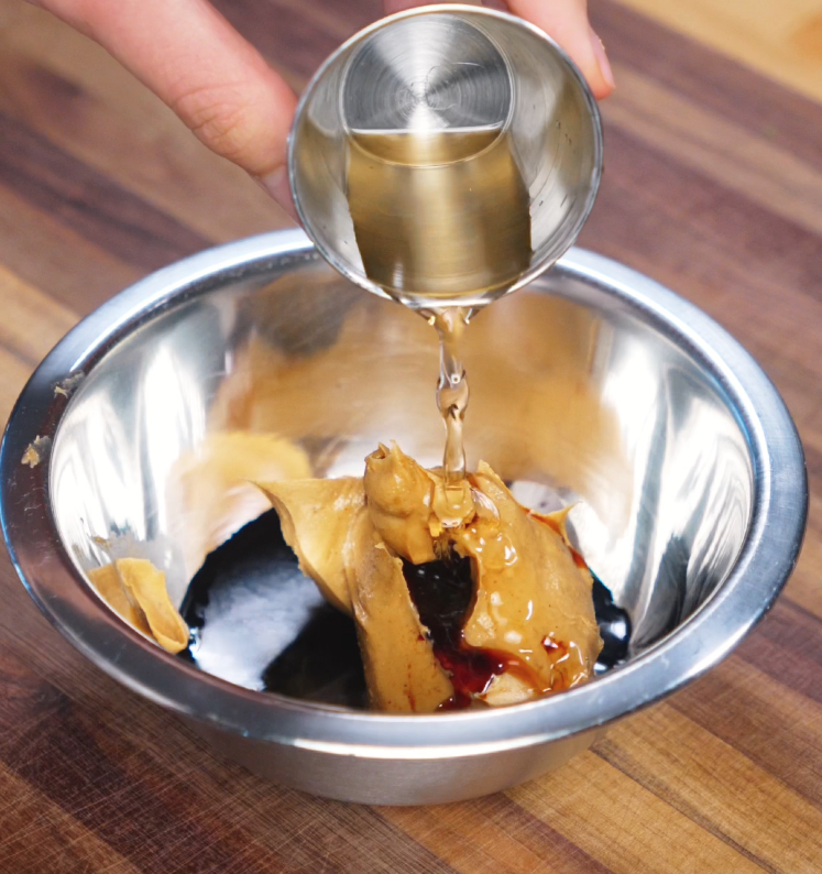 A hand pours clear vinegar from a small metal cup into a bowl containing peanut butter, soy sauces, and other marinade ingredients. The mixture is ready to be whisked into a creamy, flavorful glaze for the chicken.