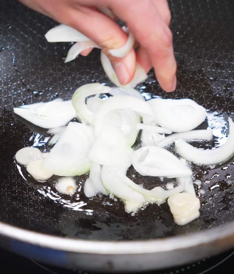 A hand holds a knife while slicing a white onion into thin half-moons on a wooden cutting board. These onions will add sweetness and depth to the savory Japanese curry broth.